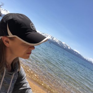 Woman sitting on the shoreline of Lake Tahoe staring out into the blue waters with snow-capped mountains in the background. She is wearing a black hat with a white rim, and you can't see her face directly.