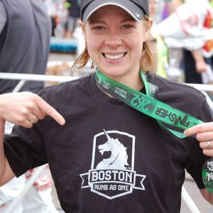 Gabe Cox at the finish line of the Boston Marathon pointing at her shirt that says Boston Runs as One and wearing a finisher medal