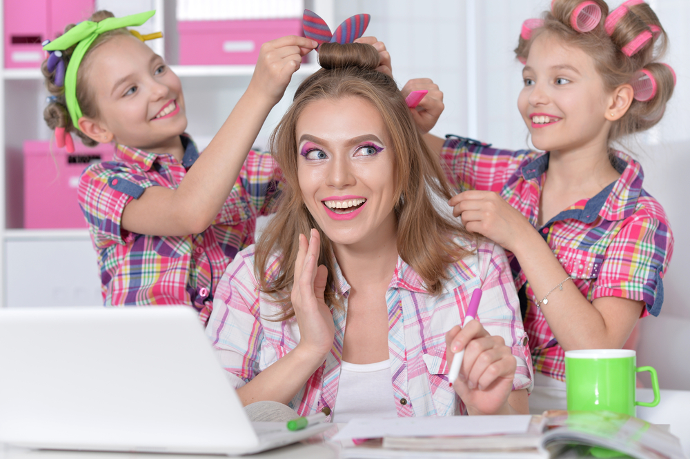 Mother sitting at a desk with her computer allowing her daughters to add curlers to her hair, all are smiling