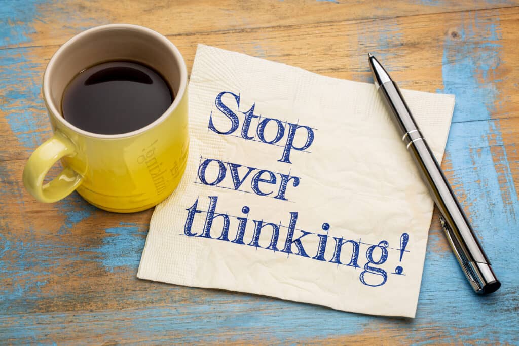 A note saying stop overthinking sitting on a wooden table with a pen lying next to it and a cup of coffee on the other side