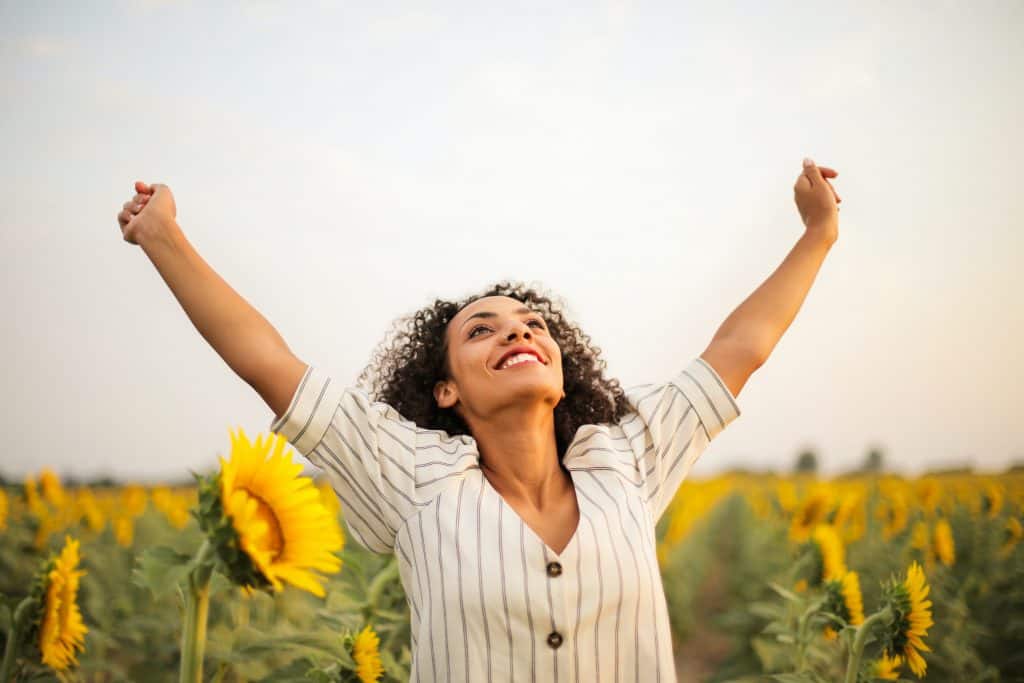 Woman with black curly hair standing in a sunflower field looking up at the sky with her arms raised in victory