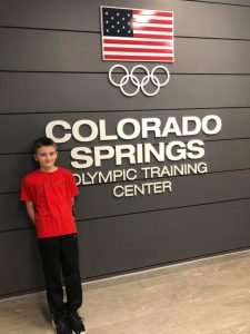 A boy standing in front of a wall that reads Colorado Springs Olympic Training Center and has an American flag an olympics logo on it