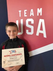 A smiling boy standing in front of a Team USA wall while holding a National Champions certificate