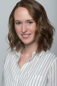 Headshot of woman with medium brown hair smiling, wearing a white and gray striped button up shirt.