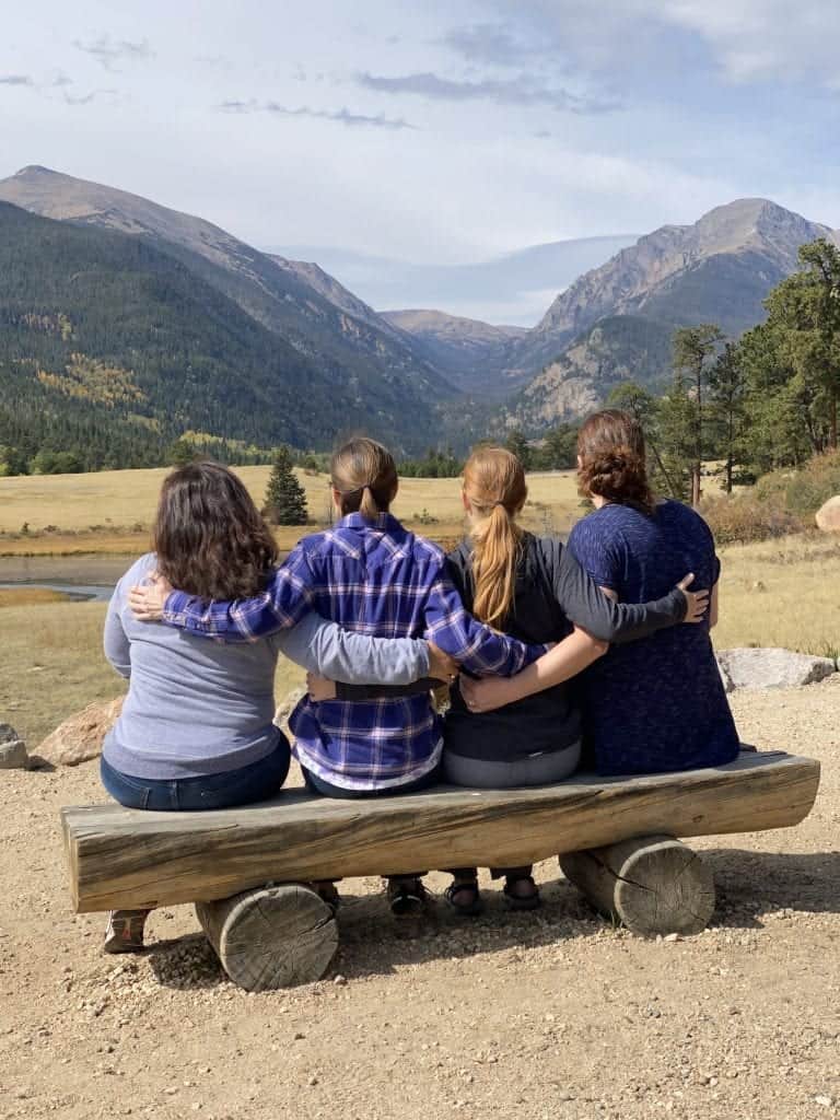 four women sitting on a bench with arms around each other staring at the rocky mountains national park.