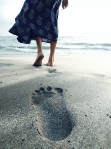 Closeup of a footprint in the sand and a woman in a dress walking away from it