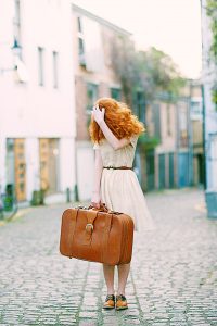 A redheaded woman standing on cobblestones holding a brown leather suitcase