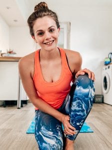 woman with her long brown hair pulled back in a bun, wearing an orange tank and blue and white workout pants. She's smiling and kneeling 