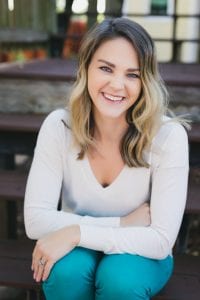 woman with dirt brown medium-length wavy hair sitting on a step outside smiling. She's wearing a white long-sleeve shirt and blue jeans.
