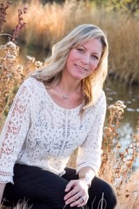 woman with medium blonde hair sitting in a field of wild flowers with a river behind her. She's wearing a white blouse with white flower designs and a silver necklace.