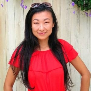 asian woman with long black hair smiling with her mouth closed in front of a light wood paneling, wearing a bright red short-sleeved shirt and sunglasses on her head.