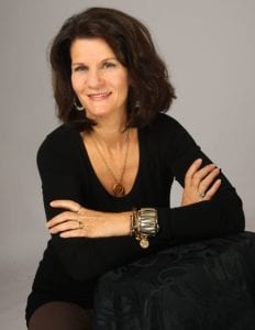 woman with medium-length dark brown hair sitting with her arms crossed and smiling. She is wearing a long-sleeve black shirt and leaning on a chair. She is wearing a long gold chain necklace with a pendant and a few gold bracelets of various sizes.