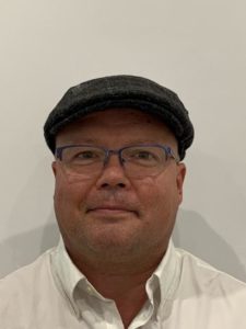 Headshot of a man wearing glasses and a hat and a white collared dress shirt