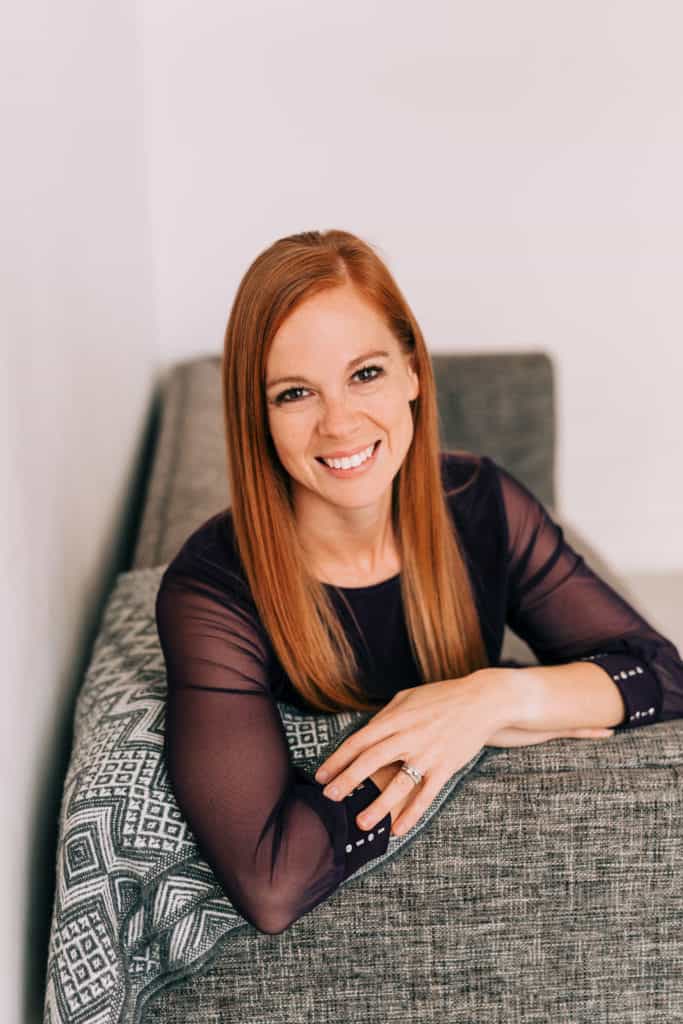 Headshot of woman with long red hair sitting on a couch with her hands folded in front of her on the armrest