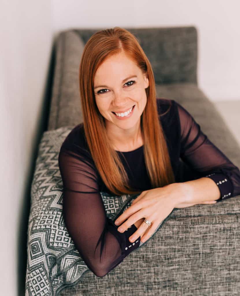 woman with long red hair smiling and leaning over the couch with her arms crossed in front of her