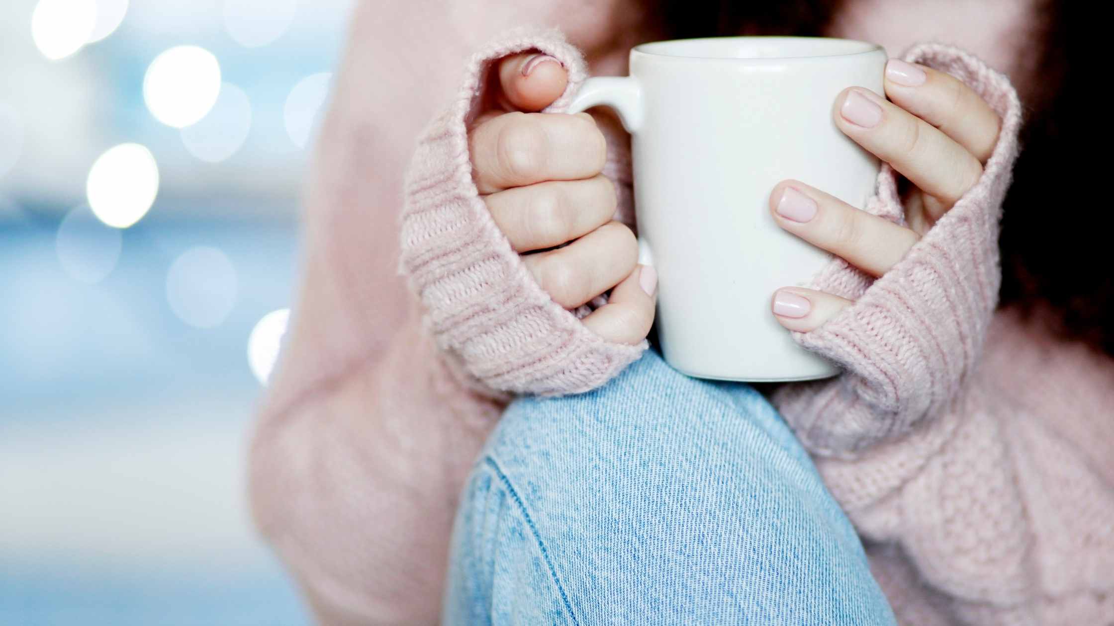 Woman sitting with a cup of coffee, just her middle body showing wearing blue jeans and pink sweater
