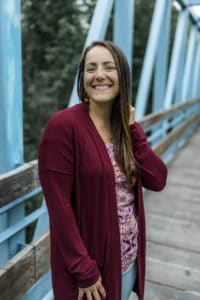 Headshot of woman with long medium hair standing on a bridge wearing a long red sweater