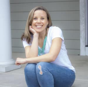 Picture of woman with medium dirt blonde hair smiling and sitting on her porch leaning her hand into the side of her face. She's wearing a white short-sleeved shirt and blue jeans.