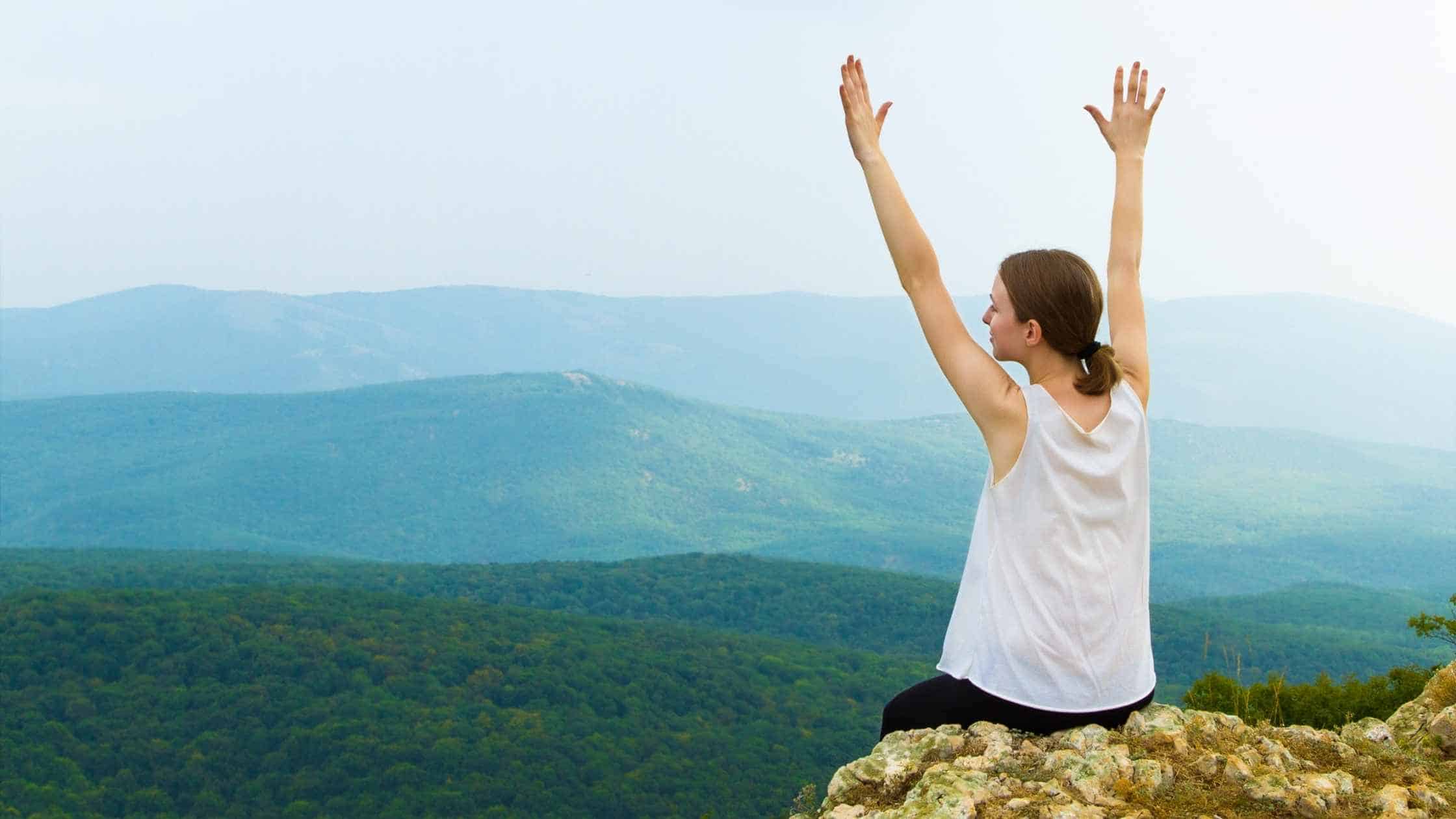 Picture of woman sitting on the edge of a mountain with her arms raised and looking away into the mountains