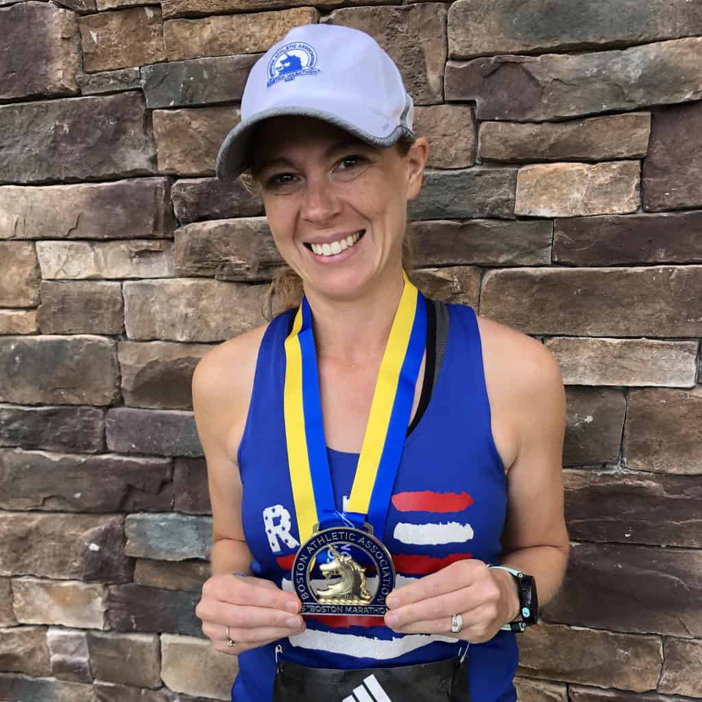 Gabe Cox standing in front of a brick wall wearing her Boston Marathon finisher's medal and holding it in her hands to show it off.