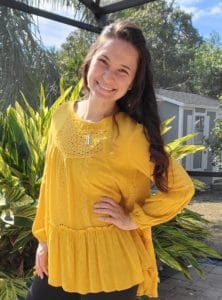 Woman with long dark brown hair wearing a yellow flowing blouse standing outside with her hand on her hip smiling. 