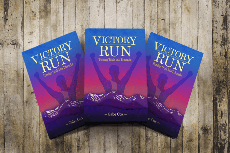 Three books laying on a wooden table, Victory Run with a cover of a sunset and snow-covered mountains with an image of a woman facing away with arms up in the air in victory