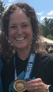 headshot of woman with medium curly brown hair smiling and holding a medal from her first ultra marathon