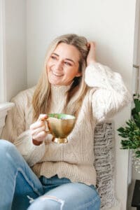 Headshot of woman with long dirt blonde, wavy hair smiling with a hand resting on her head and a cup of coffee in her other hand