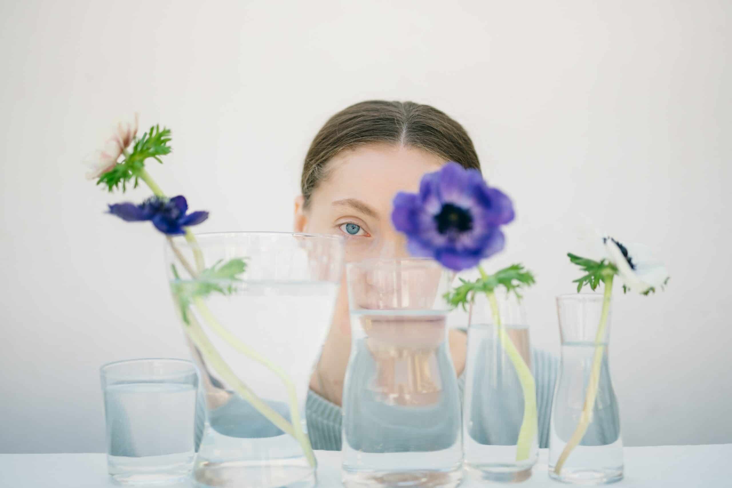 woman peeking through clear glasses of water that each have a purple flower sitting in them