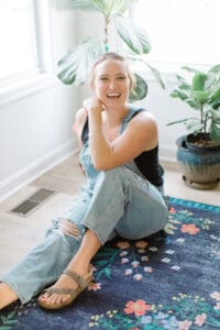 Woman sitting on the floor smiling wearing jeans and a tank with one of her knees up and an elbow resting on her knee