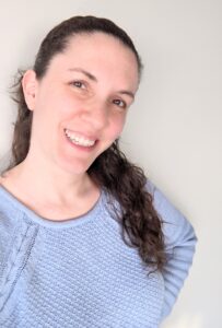 Headshot of woman with dark brown hair smiling wearing a purple sweater with hand on hip