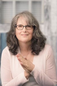 Headshot of a woman wearing a light pink sweater with brown/gray hair and glasses smiling and clasping her hands together in front of her
