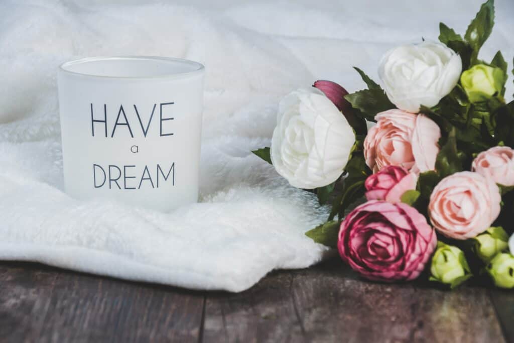 wooden table with a white fur cloth spread over it, and a mug on top of that that says "have a dream." next to that is a bouquet of pink and white roses