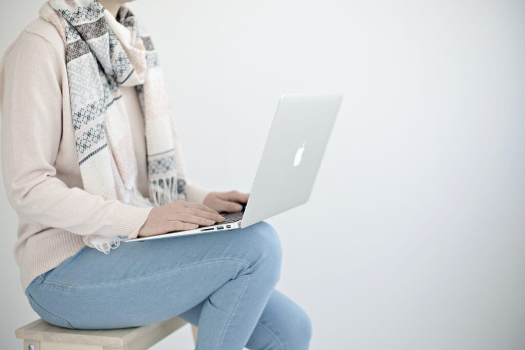 Woman sitting on a stool working on her laptop. She's wearing a light pink sweater and blue jeans