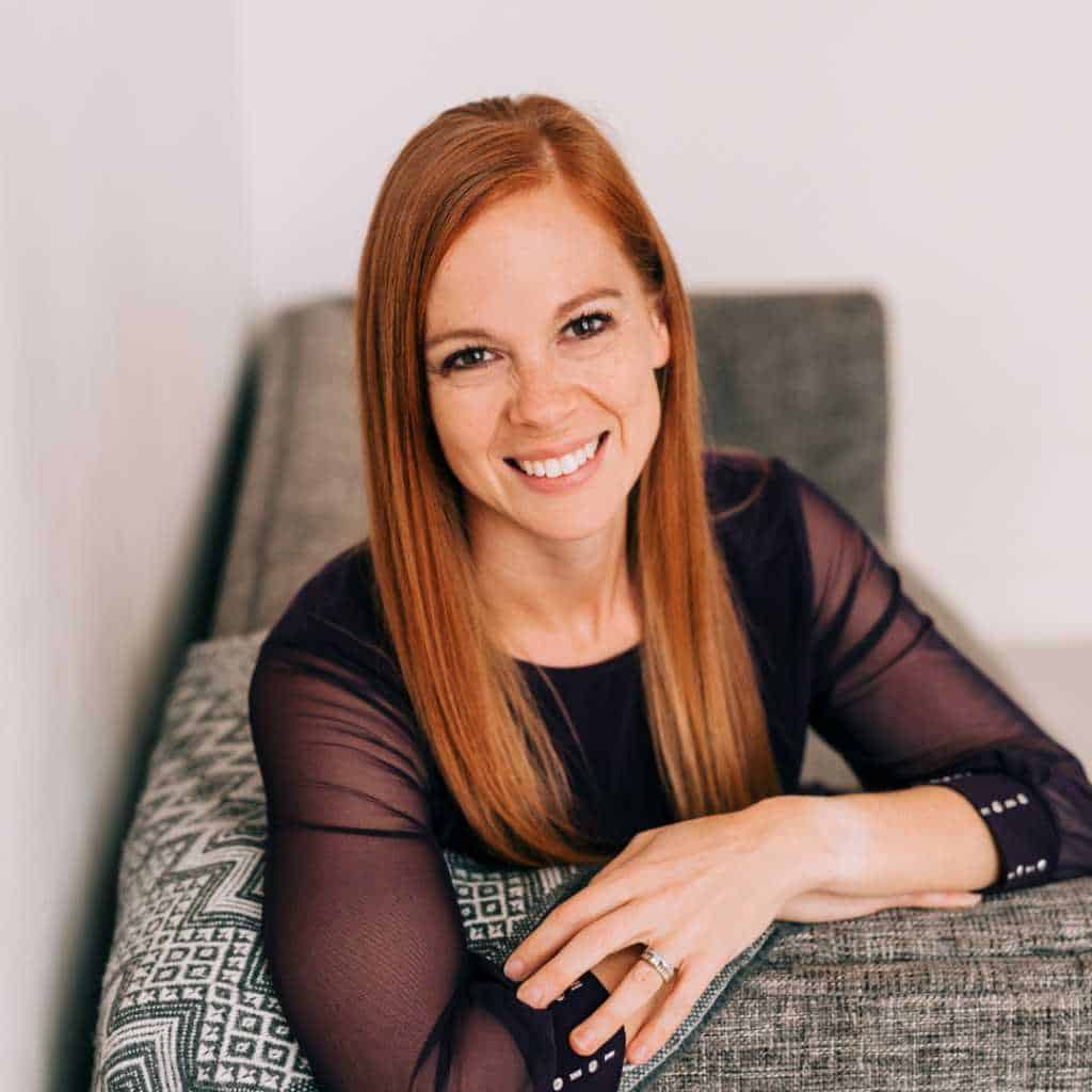 Headshot of Gabe Cox sitting on a couch and leaning over the ledge smiling. She's wearing a purple blouse