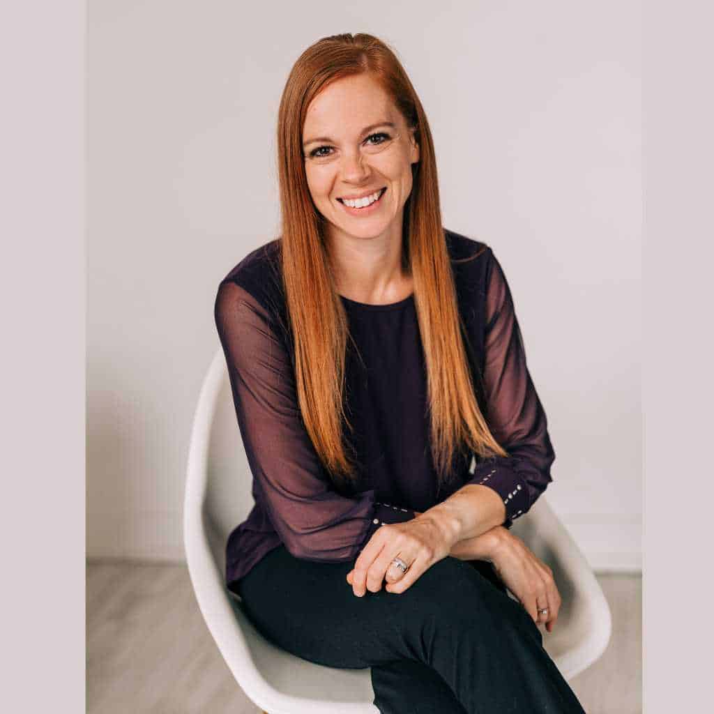 Headshot of Gabe Cox sitting in a white chair with her hands folded over her lap. She has long red hair and is wearing a purple blouse