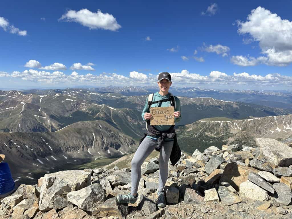 Gabe Cox standing on top of a mountain that is 14,000+ feet in elevation holding a sign saying Torrey's, which is the mountain name.