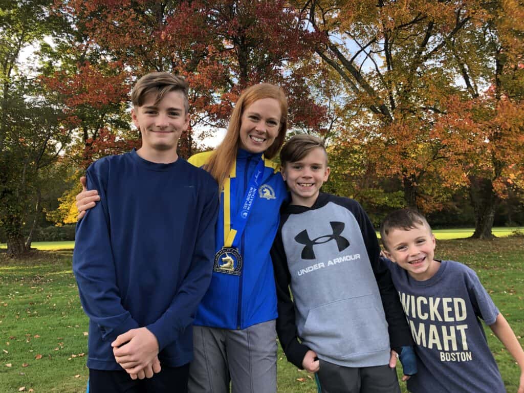 Gabe standing with her three boys outside with a background of colorful trees. She's wearing her Boston Marathon 2021 medal and jacket.