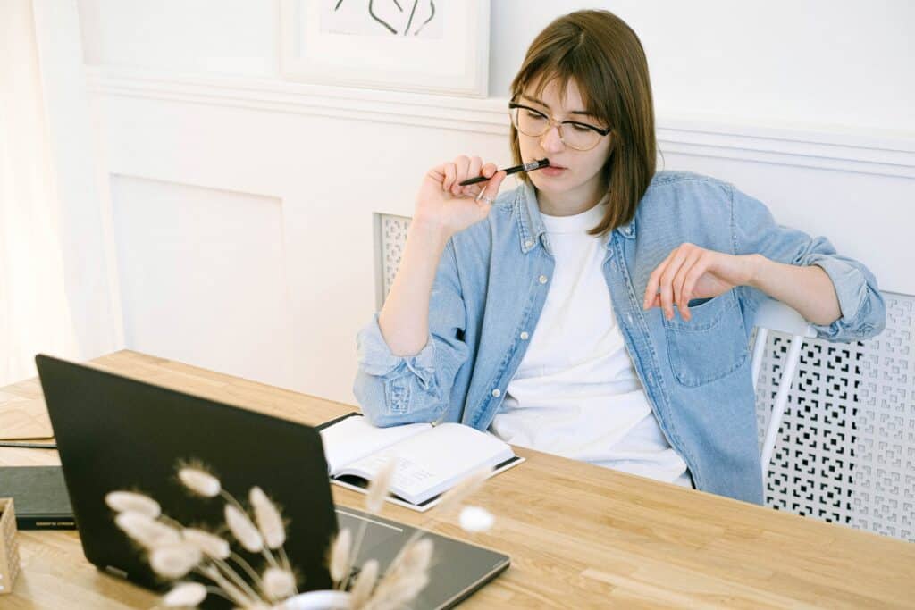 Woman with short brown hair wearing a white t-shirt and a blue button up shirt sitting at a wooden desk staring at the computer, holding a pen that is sticking out of her mouth, as if she's thinking