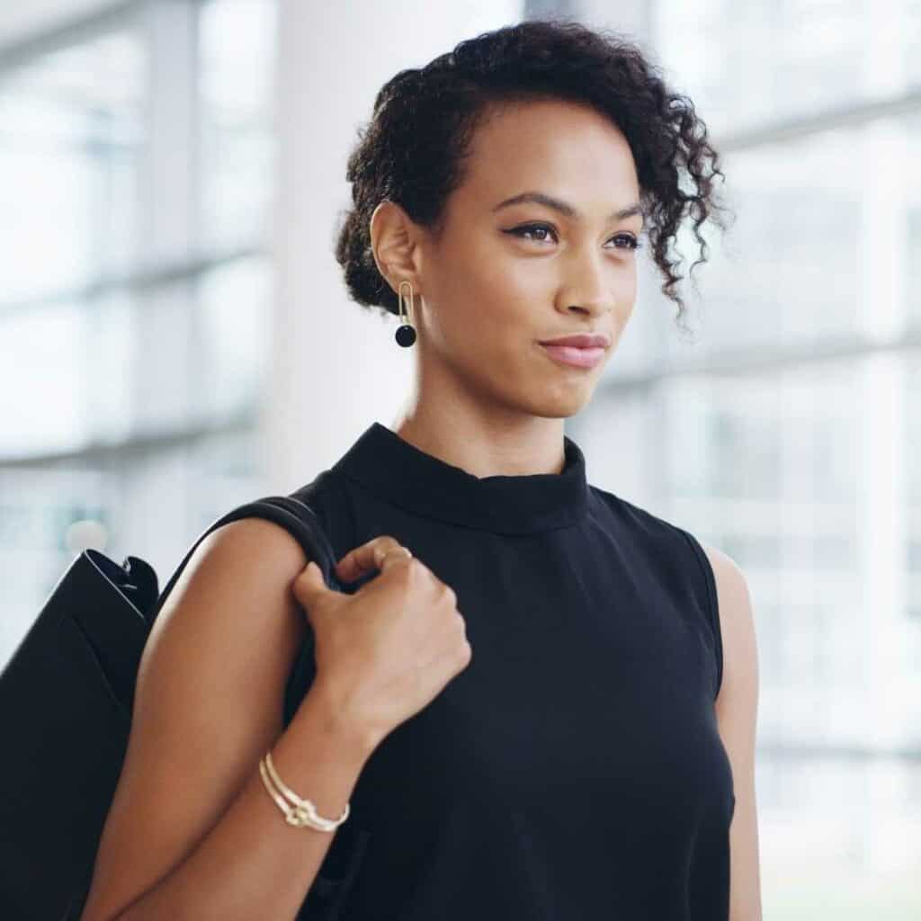 Woman with dark black curly hair pulled back standing with a bag over her shoulder looking confidently ahead as she walks.