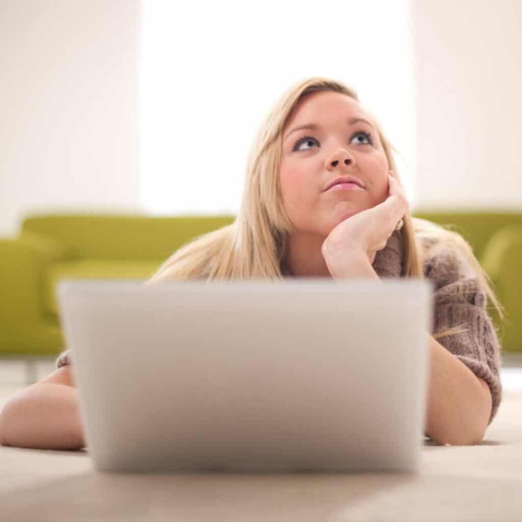 Woman with long blonde hair sitting at a table with her laptop open staring up at the sky with her hand resting on the side of her face as if she's doubting something