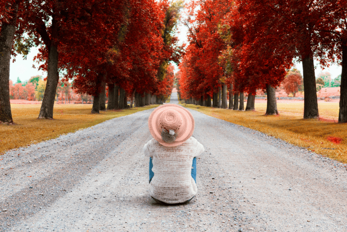 Woman sitting with back to you on the gravel road wearing a pink brimmed hat staring off into the distance of red-colored trees lining the road